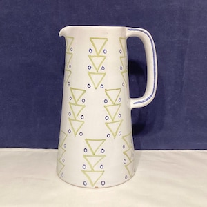 May include: White ceramic pitcher with a tall, slender shape and a handle. The pitcher is decorated with a repeating pattern of green triangles and blue dots. The handle has a blue trim.
