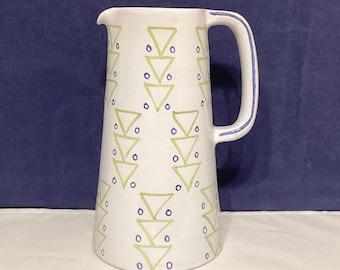 May include: White ceramic pitcher with a tall, slender shape and a handle. The pitcher is decorated with a repeating pattern of green triangles and blue dots. The handle has a blue trim.