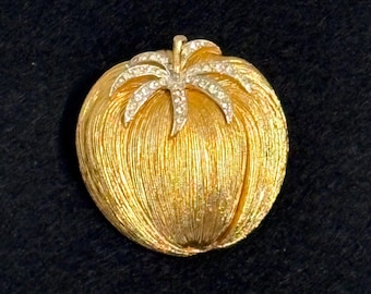 May include: A gold-toned brooch in the shape of a stylized fruit, possibly an apple or pumpkin. The brooch has a textured surface and a small, leaf-like accent at the top, embellished with small, clear crystals. The brooch is set against a dark background.