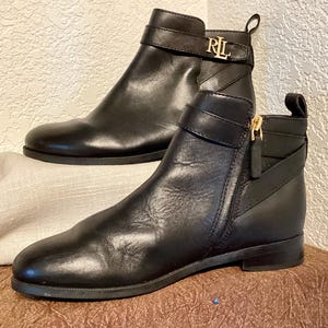 May include: A pair of black leather ankle boots. One boot features a gold buckle with the letters "RL". The other boot has a gold zipper. The boots have a low heel and a strap around the ankle.