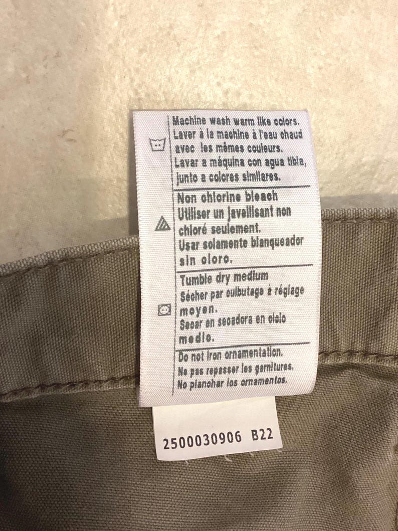 Puede incluir: Primer plano de una etiqueta de ropa con instrucciones de cuidado en varios idiomas. La etiqueta incluye instrucciones de lavado, blanqueo y secado. La etiqueta tambi&eacute;n tiene el n&uacute;mero "2500030906 B22" impreso.