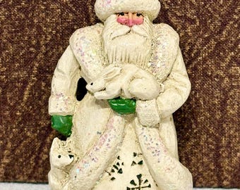 May include: A white Santa Claus figurine wearing a textured coat and hat with iridescent glitter accents. The figure holds a small white rabbit in green-gloved hands and has a small white fox standing at the base. The coat features black snowflake patterns.