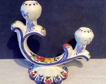 May include: A white ceramic candle holder with two candle cups. The holder is decorated with blue, red, and yellow floral patterns and a blue trim. The base is scalloped.