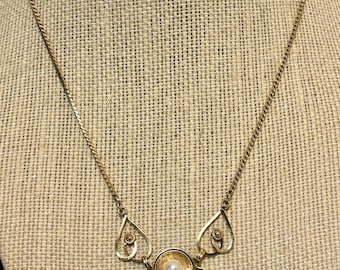 May include: A gold-tone necklace featuring a central flower-shaped pendant with a single white pearl at its core. The pendant is accented by two symmetrical teardrop-shaped side elements, all suspended from a delicate gold-tone chain link necklace.