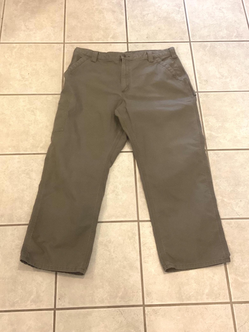 Puede incluir: Pantalones de trabajo verde oliva con m&uacute;ltiples bolsillos y dise&ntilde;o de pierna recta. Los pantalones est&aacute;n hechos de una tela duradera y est&aacute;n colocados sobre una superficie de baldosas. Cierre de bot&oacute;n y cremallera.