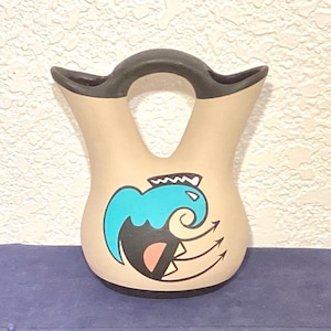 May include: Tan-colored ceramic vase with a black handle and a turquoise, black, and peach-colored design. The vase has a unique shape with two spouts and a central handle, showcasing a Southwestern-inspired aesthetic.