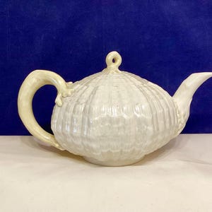 May include: A white ceramic teapot with a textured, shell-like design. The teapot has a curved handle and a spout, with a small loop on the lid. The teapot is on a white surface with a dark blue background.