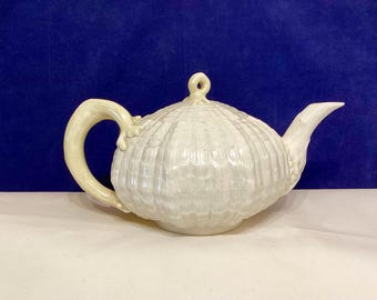 May include: A white ceramic teapot with a textured, shell-like design. The teapot has a curved handle and a spout, with a small loop on the lid. The teapot is on a white surface with a dark blue background.