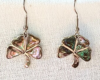 May include: A pair of silver-toned earrings featuring a shamrock design. Each earring showcases a four-leaf clover shape with iridescent abalone shell inlays, displaying hues of pink, green, and purple. The earrings have a hook closure.