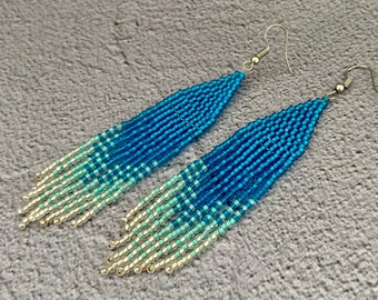 May include: A pair of beaded earrings with a gradient design. The earrings transition from shades of blue to turquoise and silver at the fringe. The earrings have silver-colored fish hook ear wires.