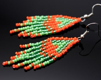 May include: A pair of beaded earrings with a fringe design. The earrings feature a pattern of orange and green beads, with silver-colored ear wires. The earrings are displayed against a black background.