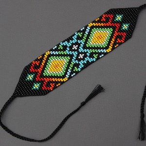 May include: A beaded bracelet featuring a black background and a central geometric pattern in yellow, green, blue, and red. It includes a black braided cord and tassel. This accessory is suitable for various occasions.
