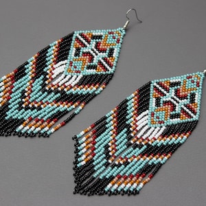 Pode incluir: Um par de brincos de contas com um design geom&eacute;trico. Os brincos apresentam uma parte superior quadrada com um padr&atilde;o de contas turquesa, pretas, brancas, vermelhas e laranja. Longas franjas pendem da parte inferior, com contas pretas, turquesas e brancas.