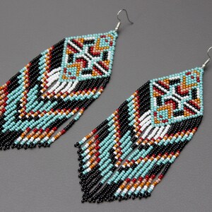 Pode incluir: Um par de brincos de contas com um design geom&eacute;trico. Os brincos apresentam um topo em forma de diamante com um padr&atilde;o de contas turquesa, pretas, brancas, vermelhas e laranja. Longas franjas pretas pendem da parte inferior.