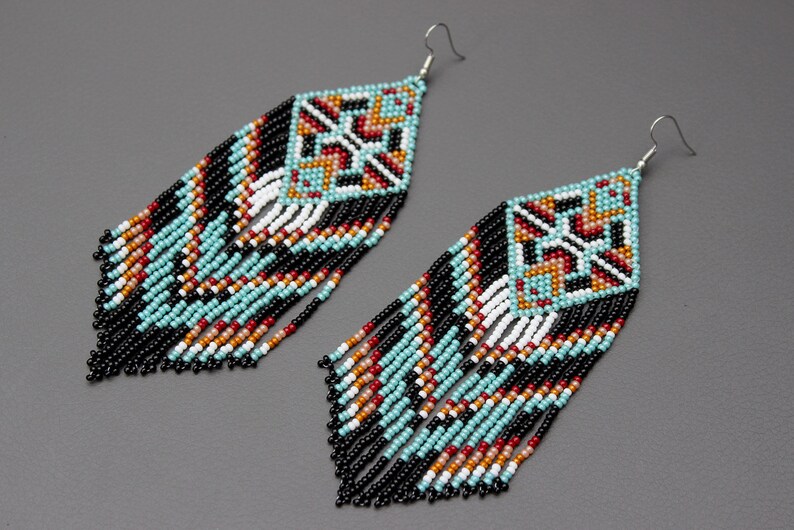 Pode incluir: Um par de brincos de contas com um design geom&eacute;trico. Os brincos apresentam um topo em forma de diamante com um padr&atilde;o de contas turquesa, pretas, brancas, vermelhas e laranja. Longas franjas pretas pendem da parte inferior.