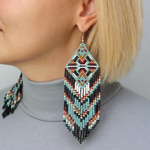 Pode incluir: Brincos em forma de diamante com um padr&atilde;o geom&eacute;trico em turquesa, preto, vermelho, branco e castanho. Os brincos t&ecirc;m uma franja de contas pretas e s&atilde;o suspensos por ganchos de prata. Um acess&oacute;rio marcante.