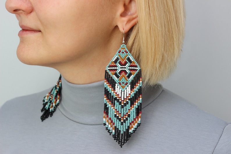 Pode incluir: Brincos em forma de diamante com um padr&atilde;o geom&eacute;trico em turquesa, preto, vermelho, branco e castanho. Os brincos t&ecirc;m uma franja de contas pretas e s&atilde;o suspensos por ganchos de prata. Um acess&oacute;rio marcante.