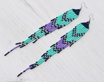 May include: A pair of long beaded earrings in turquoise, purple, black, and silver. The earrings have a geometric pattern and dangle from silver-colored hooks. The beads are arranged in a fringe design.