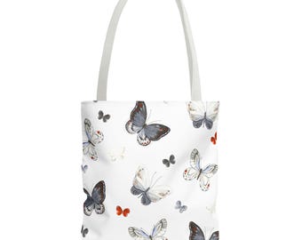 May include: A white tote bag featuring a repeating pattern of watercolour-style butterflies in shades of grey, black, and orange. The bag has two long white shoulder straps and a rectangular body with a clean, simple design for carrying items.