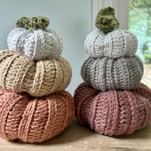May include: A stack of three crocheted pumpkins in shades of beige, gray, and pink. The pumpkins are decorated with green leaves on top.