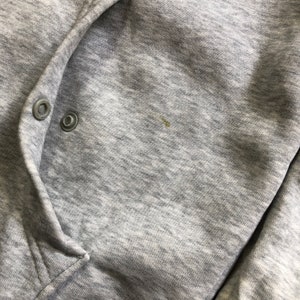 K&ouml;nnte beinhalten: Nahaufnahme eines grauen Kapuzenpullovers mit zwei silbernen Druckkn&ouml;pfen. Der Stoff ist ein weiches, meliertes Material. Ein kleiner Fleck ist auf der rechten Seite des Kleidungsst&uuml;cks sichtbar.