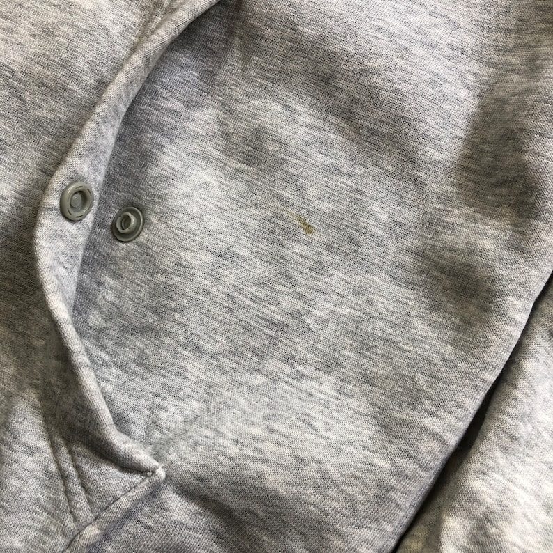 K&ouml;nnte beinhalten: Nahaufnahme eines grauen Kapuzenpullovers mit zwei silbernen Druckkn&ouml;pfen. Der Stoff ist ein weiches, meliertes Material. Ein kleiner Fleck ist auf der rechten Seite des Kleidungsst&uuml;cks sichtbar.