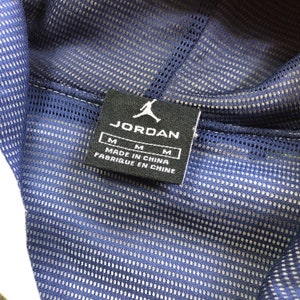 K&ouml;nnte beinhalten: Nahaufnahme eines marineblauen Mesh-Stoffs mit wei&szlig;em Futter. Ein schwarzes Etikett mit dem wei&szlig;en "JORDAN"-Logo und dem Text "MADE IN CHINA FABRIQUE EN CHINE" ist auf den Stoff gen&auml;ht. Das Etikett hat auch die Gr&ouml;&szlig;e "M".