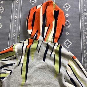 K&ouml;nnte beinhalten: Ein grauer Kapuzenpullover mit einem farbenfrohen, abstrakten Muster. Die Kapuze hat ein orangefarbenes, wei&szlig;es und marineblaues Design, w&auml;hrend der K&ouml;rper gr&uuml;ne, marineblaue und wei&szlig;e Akzente aufweist. Der Kapuzenpullover ist aus weichem Material.