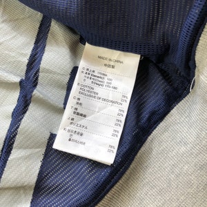 K&ouml;nnte beinhalten: Nahaufnahme eines Kleidungsst&uuml;cketiketts mit Text in mehreren Sprachen, darunter Englisch und Chinesisch. Das Etikett listet die Stoffzusammensetzung auf: Baumwolle und Polyester. Das Kleidungsst&uuml;ck hat eine blaue Mesh-Verzierung und einen hellgrauen Stoff.