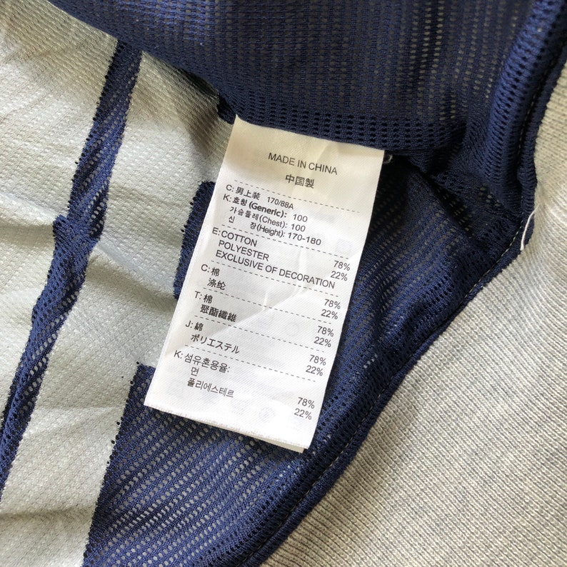 K&ouml;nnte beinhalten: Nahaufnahme eines Kleidungsst&uuml;cketiketts mit Text in mehreren Sprachen, darunter Englisch und Chinesisch. Das Etikett listet die Stoffzusammensetzung auf: Baumwolle und Polyester. Das Kleidungsst&uuml;ck hat eine blaue Mesh-Verzierung und einen hellgrauen Stoff.