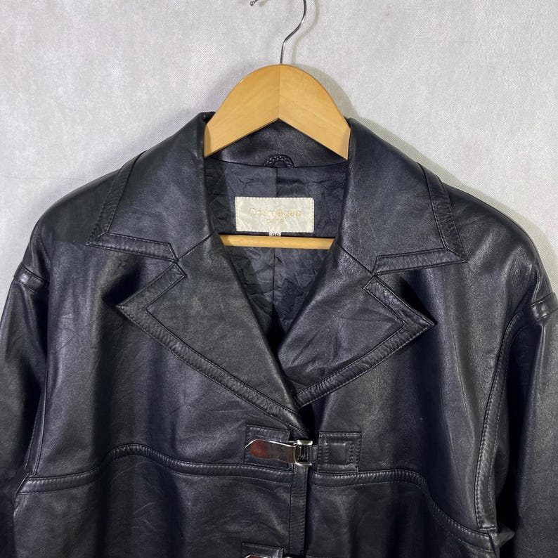 May include: Black leather jacket with a buckle closure and a collar. The jacket has a  stitched design and a label that reads "Genuine Leather".