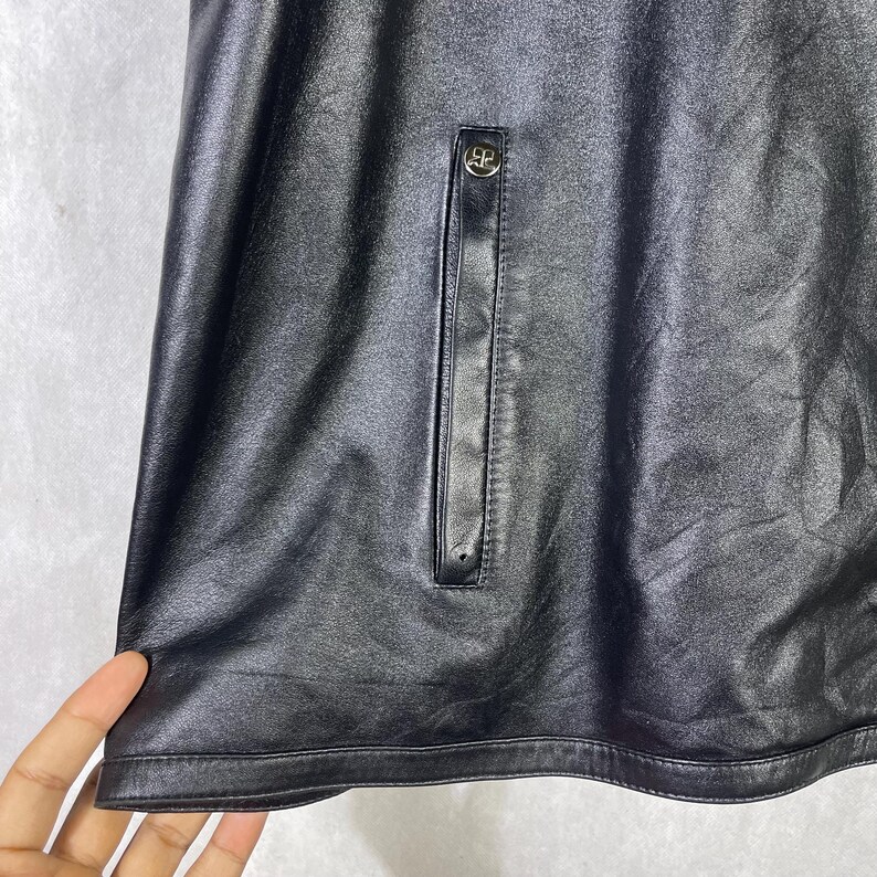 May include: Black leather jacket with a zippered pocket on the front. The pocket has a silver zipper pull with a small metal logo.