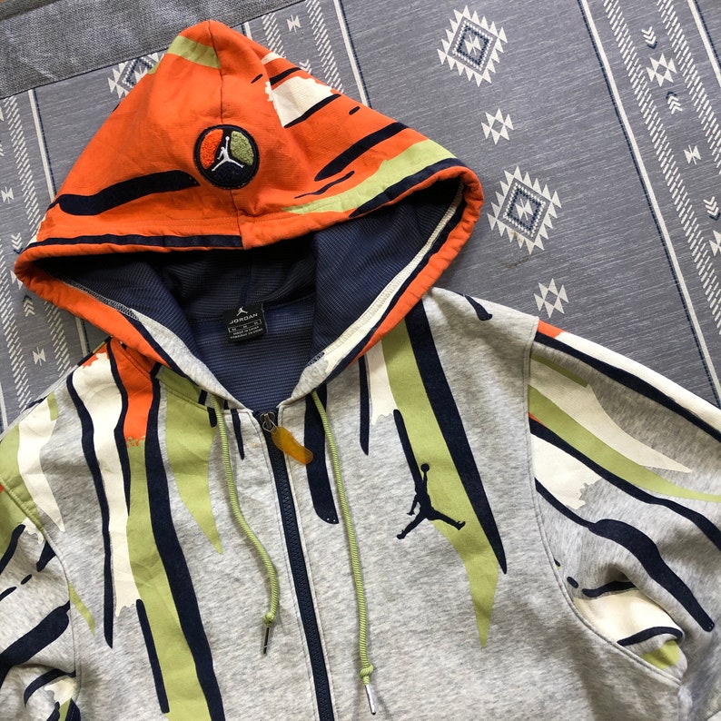 K&ouml;nnte beinhalten: Ein grauer Kapuzenpullover mit Rei&szlig;verschluss und einem farbenfrohen Muster und einer Kapuze. Die Kapuze ist orange mit schwarzen und wei&szlig;en Streifen und einem kreisf&ouml;rmigen Logo. Der K&ouml;rper des Hoodies hat gr&uuml;ne, wei&szlig;e und marineblaue Streifen. Ein schwarzes Jumpman-Logo befindet sich auf der Brust.