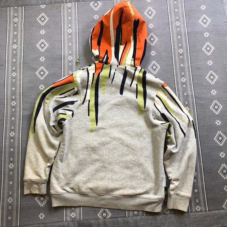K&ouml;nnte beinhalten: Ein grauer Kapuzenpullover mit einem farbenfrohen abstrakten Design auf der Kapuze und dem oberen R&uuml;cken. Das Design zeigt orange, gr&uuml;ne und schwarze vertikale Streifen. Der Kapuzenpullover ist aus einem weichen, bequemen Material.