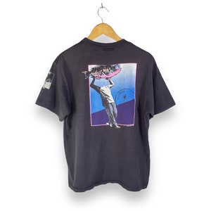 May include: Black t-shirt with a graphic print of a person holding a fish. The graphic is in a blue, pink, and purple color scheme. The text "PALMILLA" is printed on the graphic.