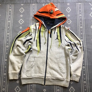 K&ouml;nnte beinhalten: Ein grauer Kapuzenpullover mit Rei&szlig;verschluss und farbenfrohem Design. Die Kapuze ist orange mit einem kreisf&ouml;rmigen Logo. Die &Auml;rmel und die Vorderseite weisen gr&uuml;ne, schwarze und wei&szlig;e Streifen auf. Ein schwarzes Jumpman-Logo befindet sich auf der Brust.