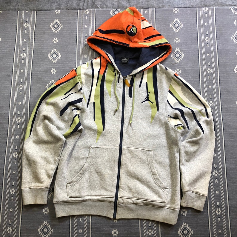 K&ouml;nnte beinhalten: Ein grauer Kapuzenpullover mit Rei&szlig;verschluss und farbenfrohem Design. Die Kapuze ist orange mit einem kreisf&ouml;rmigen Logo. Die &Auml;rmel und die Vorderseite weisen gr&uuml;ne, schwarze und wei&szlig;e Streifen auf. Ein schwarzes Jumpman-Logo befindet sich auf der Brust.