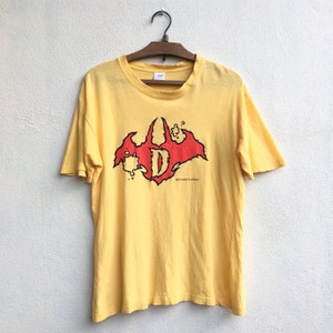 May include: Yellow t-shirt with a red graphic of a stylized "D" inside a bat-like shape. The graphic has a distressed look and says "DYNAMIC PLUNDER" at the bottom.