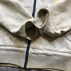 K&ouml;nnte beinhalten: Nahaufnahme eines grauen Kapuzenpullovers mit Rei&szlig;verschluss und marineblauen B&uuml;ndchen. Die B&uuml;ndchen sind umgeschlagen. Der Hoodie besteht aus einem weichen, bequemen Material.