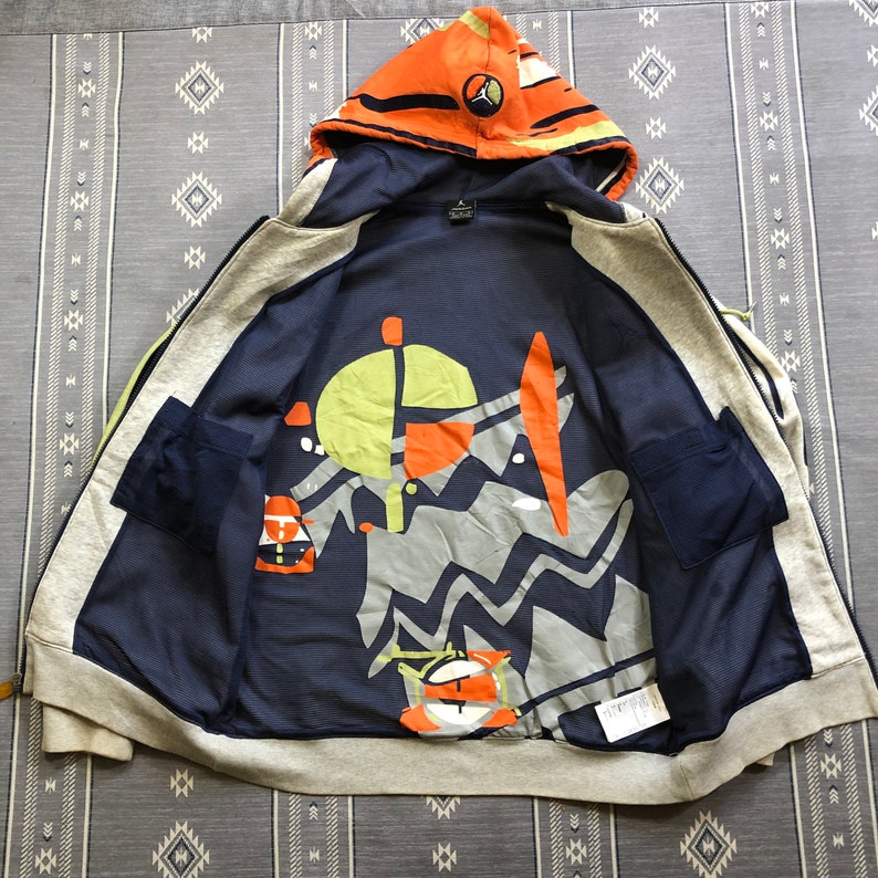 K&ouml;nnte beinhalten: Offener Kapuzenpullover mit Rei&szlig;verschluss und marineblauem Innenfutter mit farbenfrohem, abstraktem Design. Die Kapuze ist orange mit schwarzen Streifen und einem Logo. Die Au&szlig;enseite ist grau mit marineblauen Akzenten und Taschen.