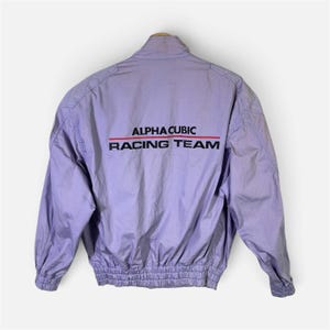 May include: A light purple jacket with a high collar, long sleeves, and an elasticized waistband. The back features the text ALPHA CUBIC RACING TEAM in black lettering, separated by a thin horizontal red line across the upper back panel.