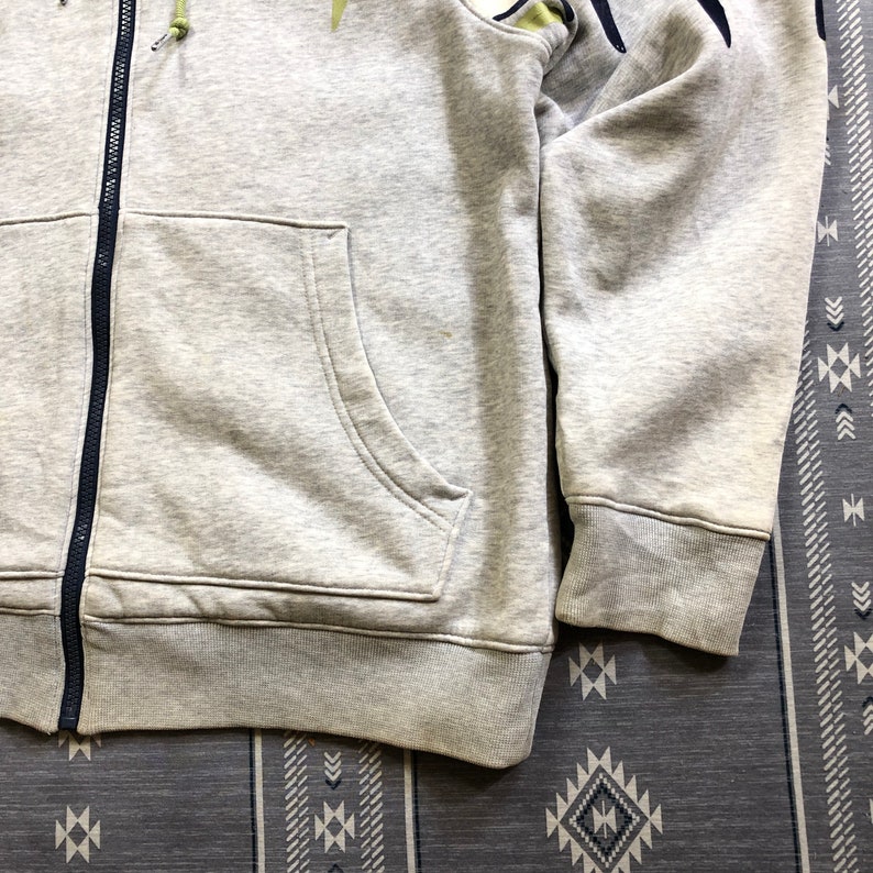 K&ouml;nnte beinhalten: Ein hellgrauer Kapuzenpullover mit Rei&szlig;verschluss, Vordertasche und marineblauem Rei&szlig;verschluss. Der Hoodie hat einen gerippten Saum und B&uuml;ndchen. Die &Auml;rmel haben ein dunkelblaues und gr&uuml;nes Design am Oberarm.