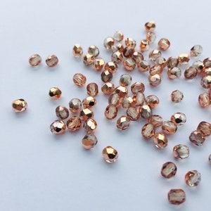 May include: A collection of small, faceted glass beads in a rose gold colour. The beads are scattered on a white surface.