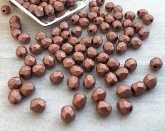 May include: A scattering of copper-coloured faceted glass beads, some in a small metal scoop. The beads are small and have a rough, textured surface.