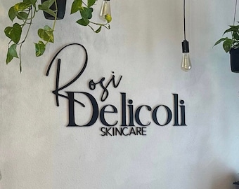 May include: A white ceiling with a crystal chandelier and four hanging light bulbs. The wall is white with a black sign that reads "Rosi Delicoli Skincare". There are three hanging plants with green leaves.