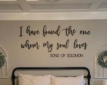 Puede incluir: Un dormitorio con un edredón a rayas blancas y marrones sobre un marco de cama de metal negro. La pared detrás de la cama tiene una calcomanía de vinilo negro con el texto "I have found the one whom my soul loves Song of Solomon". Hay dos arcos decorativos blancos con vegetación a cada lado de la cama.