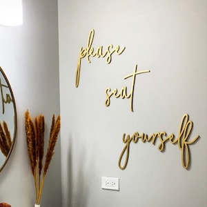May include: Gold cursive lettering on a white wall that reads "please seat yourself". The lettering is mounted on the wall in a bathroom with a white sink, a gold faucet, and a white towel.