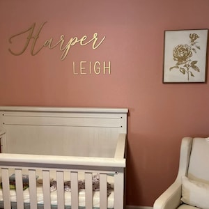 May include: A white crib with a white mattress and a pink wall with gold lettering that says "Harper Leigh" and a framed picture of a gold rose on a white background.