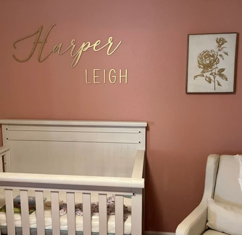 May include: A white crib with a white mattress and a pink wall with gold lettering that says "Harper Leigh" and a framed picture of a gold rose on a white background.