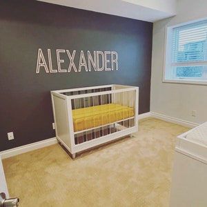 May include: A white crib with a yellow mattress and a white wooden name sign that says "ALEXANDER" on a dark gray wall.